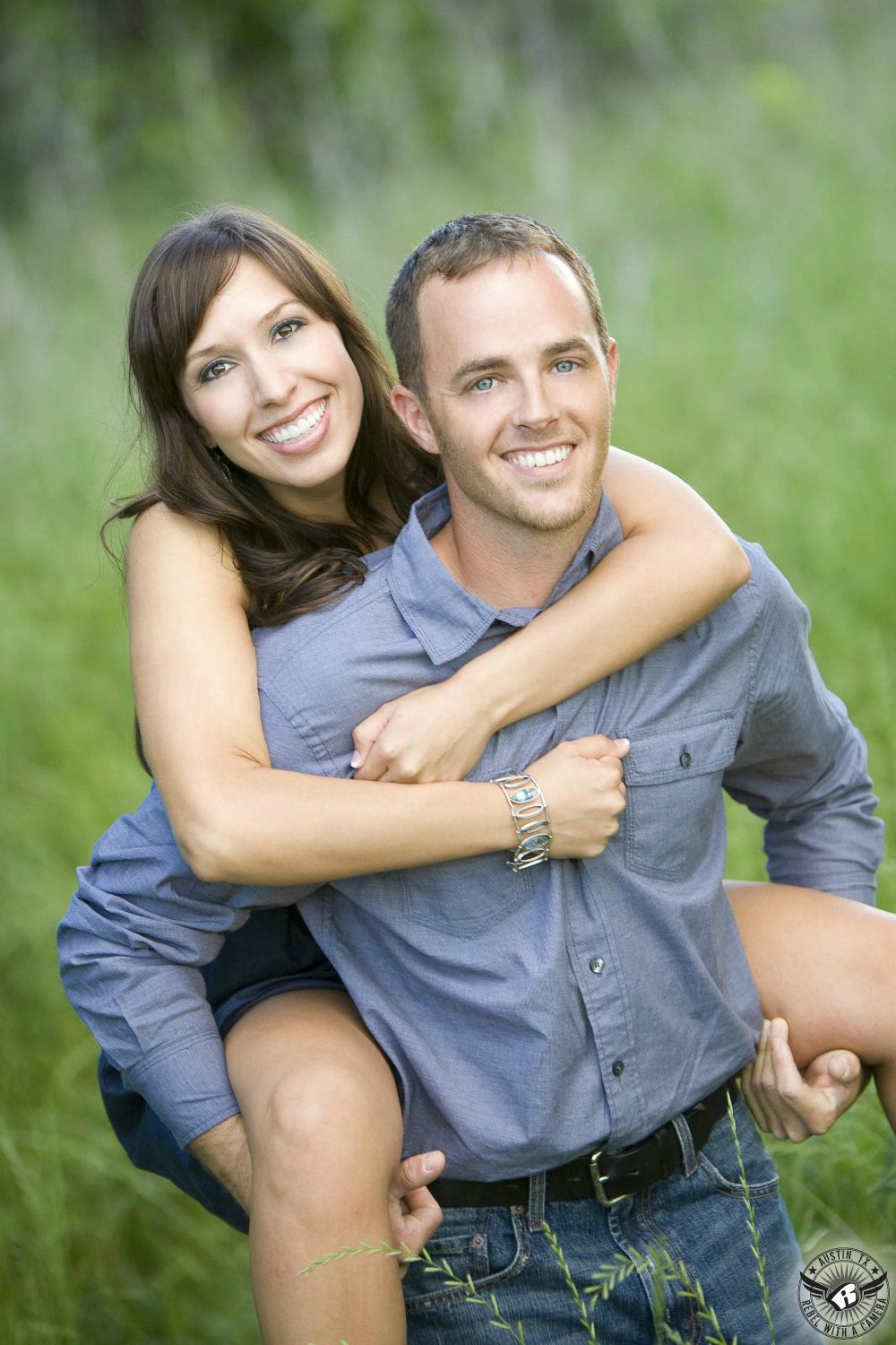 dark haired happy couple in trendy grey and blue clothes piggy back through tall green grass in taylor texas 